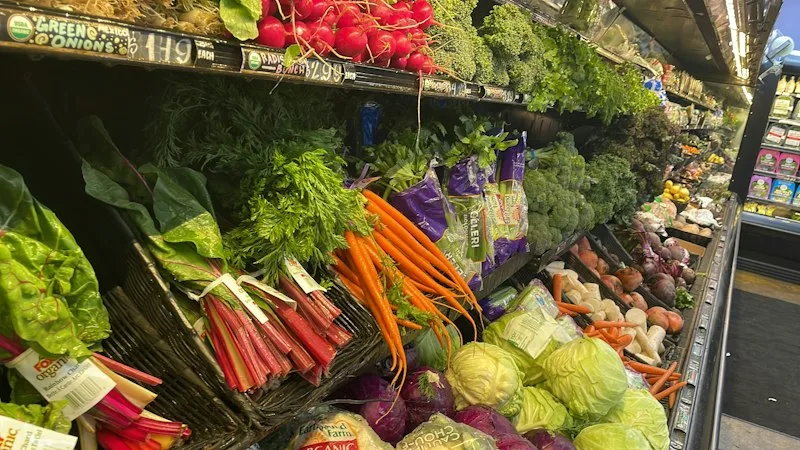 A produce aisle in a grocery co-op, with rainbow chard, carrots and cabbages in slanted baskets and price signs written in chalk above them.
