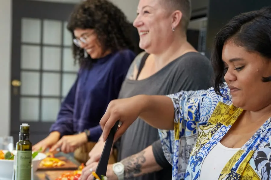 Three women laughing as they cook side by side at a kitchen counter, with chopped peppers on the boards in front of them.