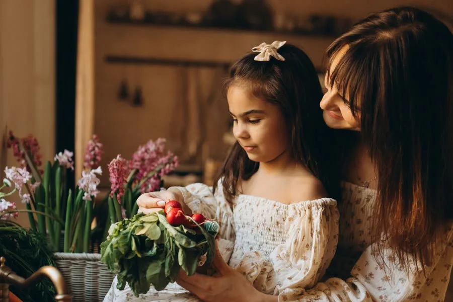 A mother and her young daughter holding a bunch of radishes and greens by a kitchen window, with carrots and spring flowers beside them.