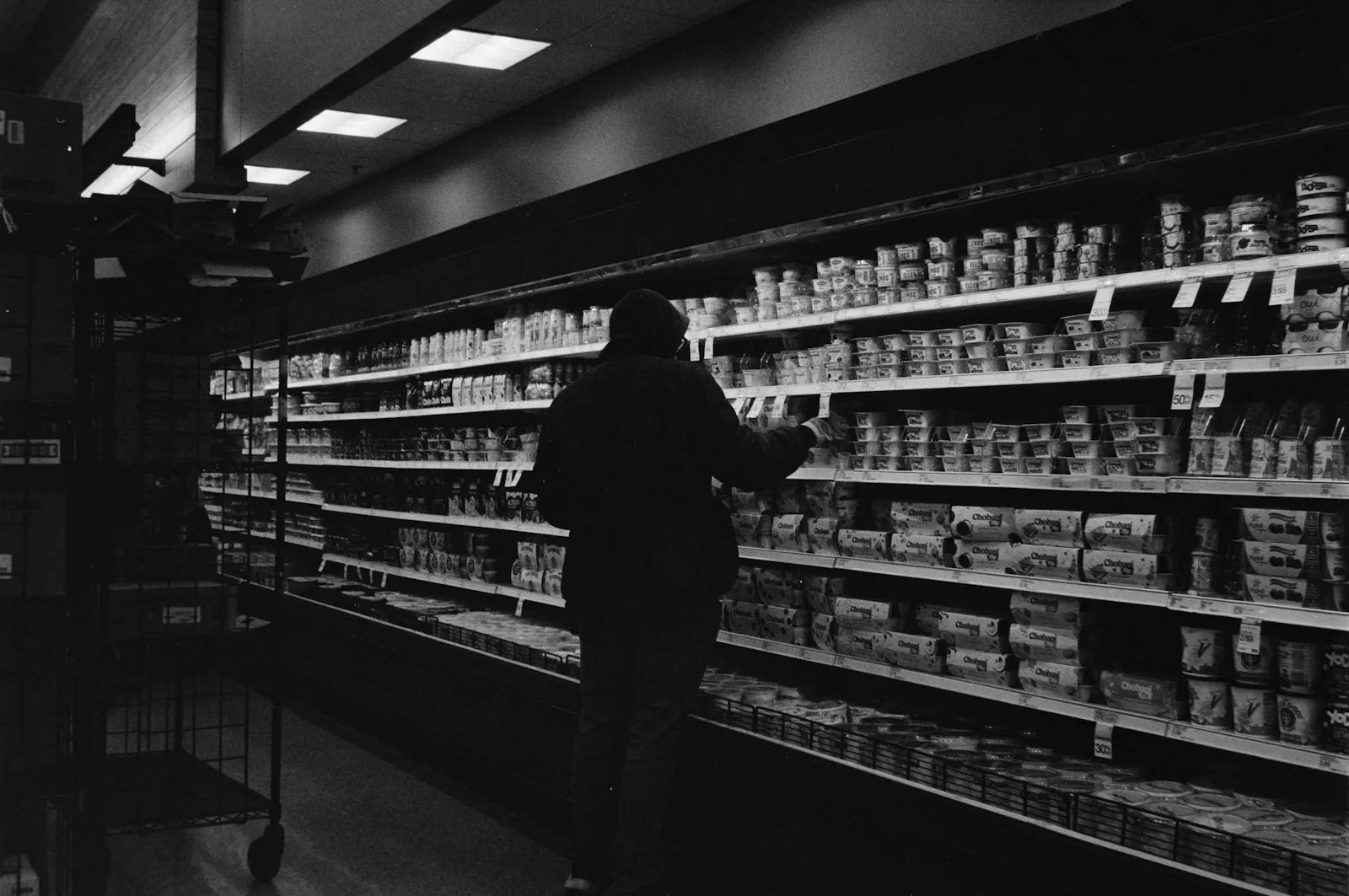 A shopper reaching for a tub on a long refrigerated dairy shelf, photographed in black and white.