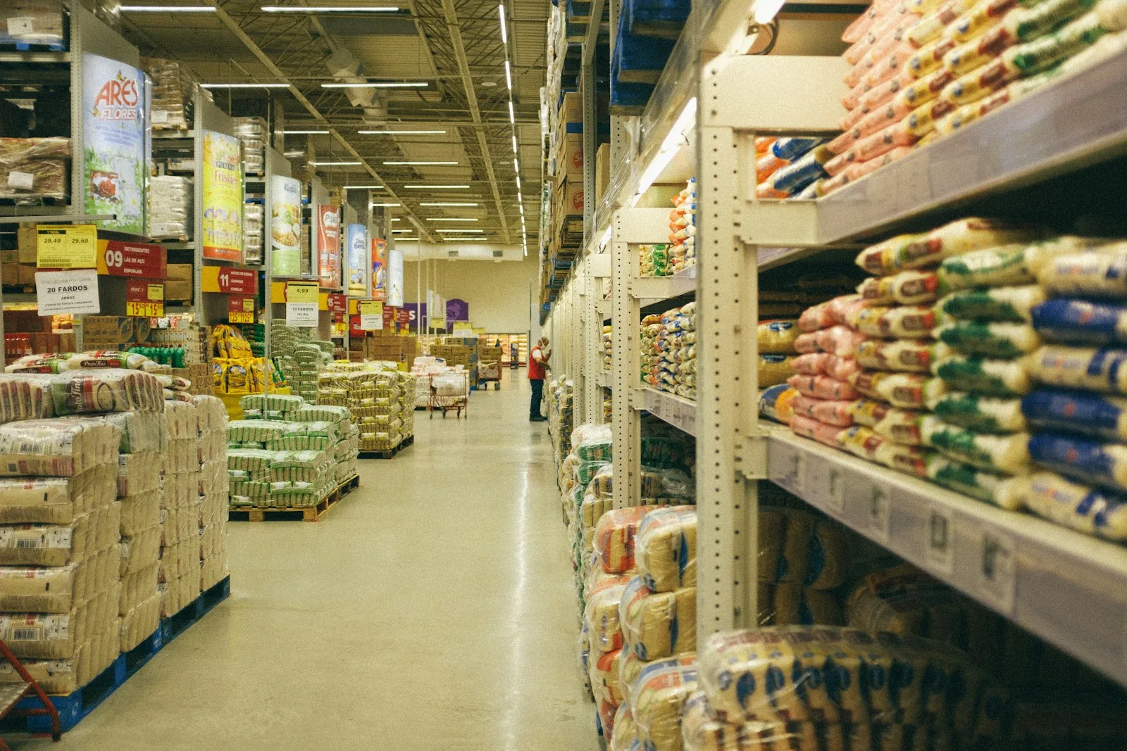 A wide aisle in a warehouse store, with bulk bags of rice and grain stacked on pallets and on tall shelving.
