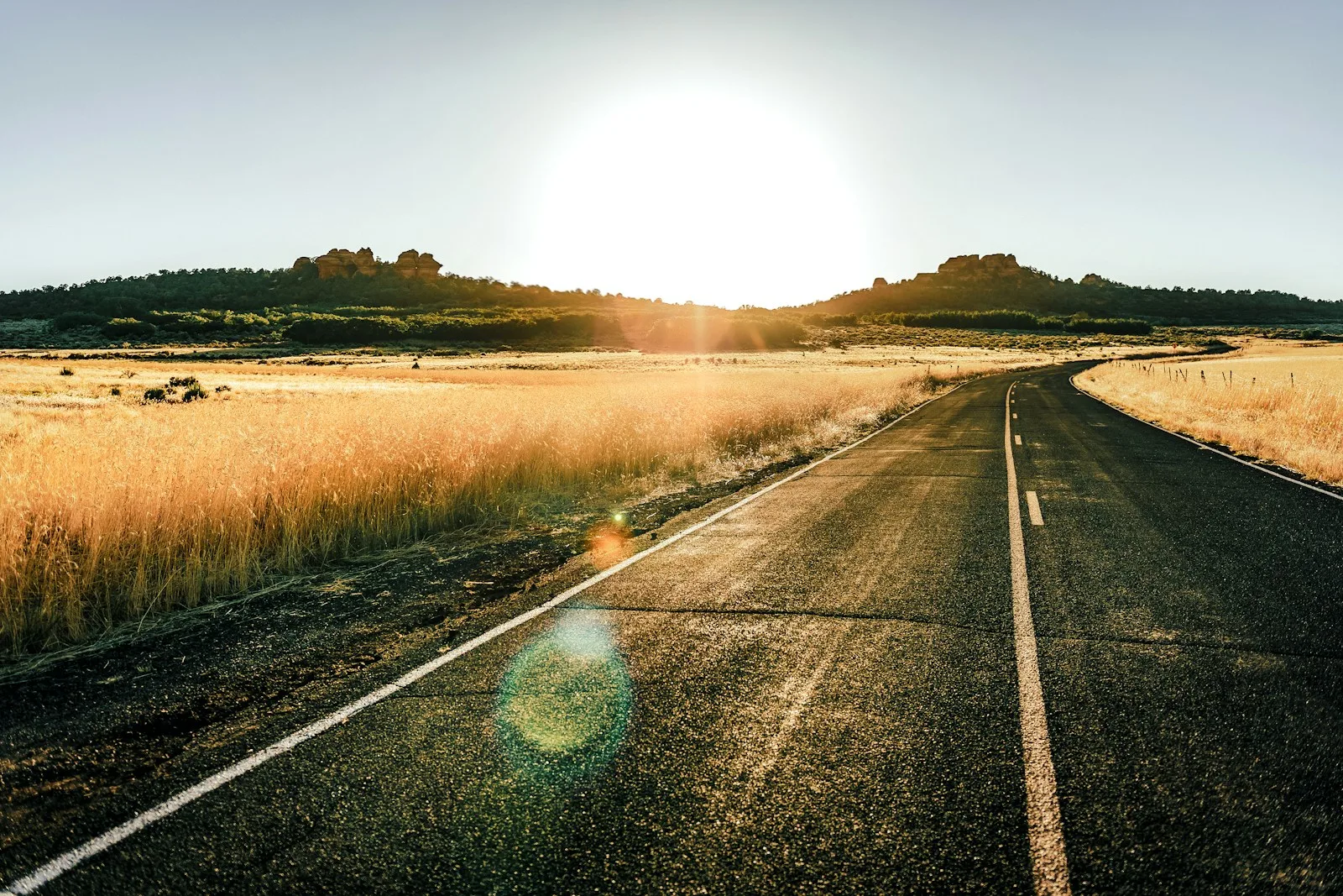 An empty two-lane road running through dry golden fields toward low hills at sunrise.