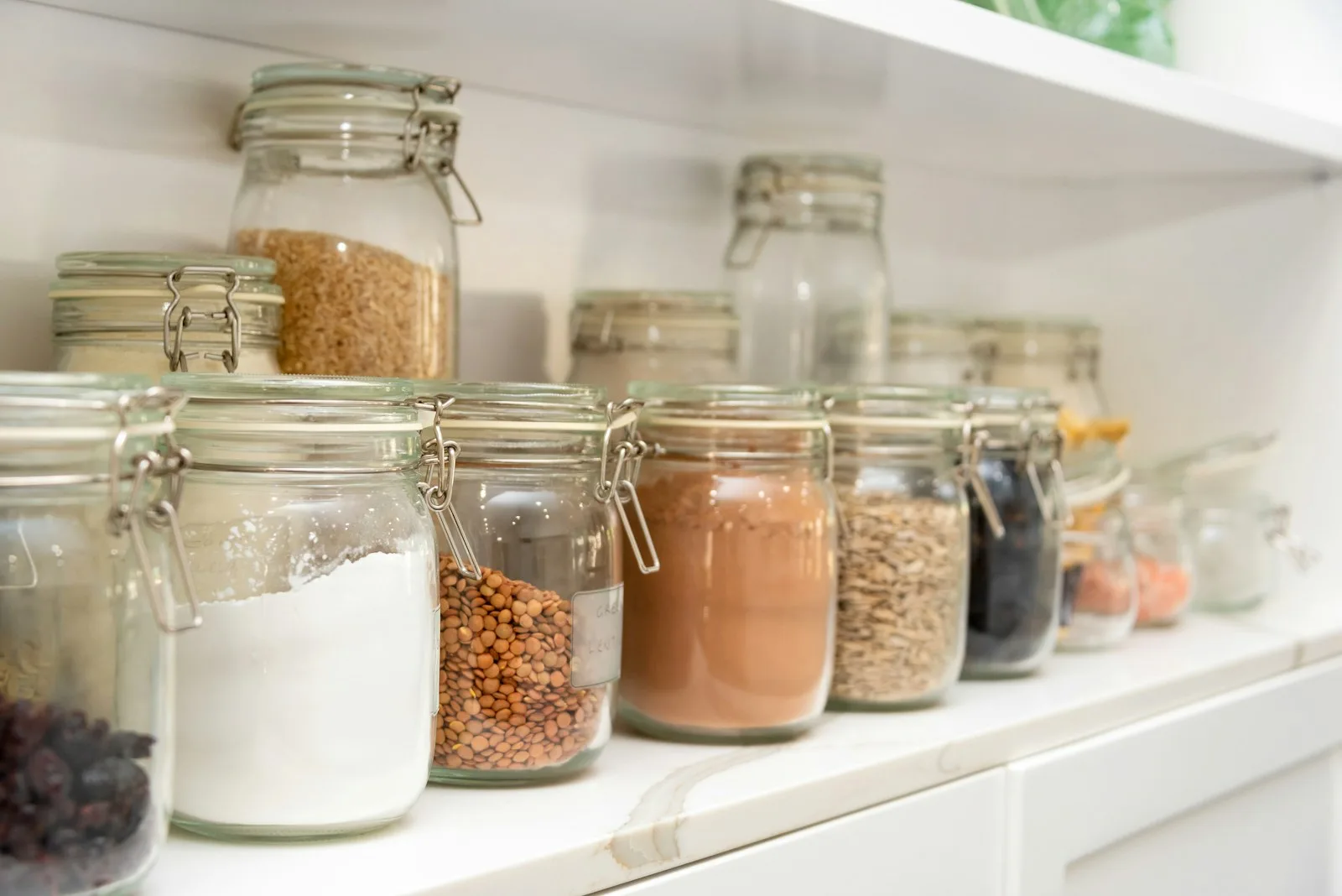Glass jars of flour, lentils, oats and other dry goods lined up on a white pantry shelf.